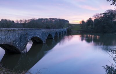 a pink purple glow with the eight arch bridge leading to the sun just beginning to peep out over the fields at stackpole