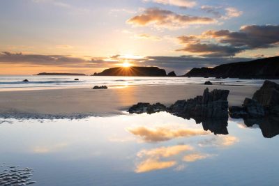 a sun burst ray effect as the sun sets over a moody blue marloes beach with orange clouds reflected in a calm rockpool